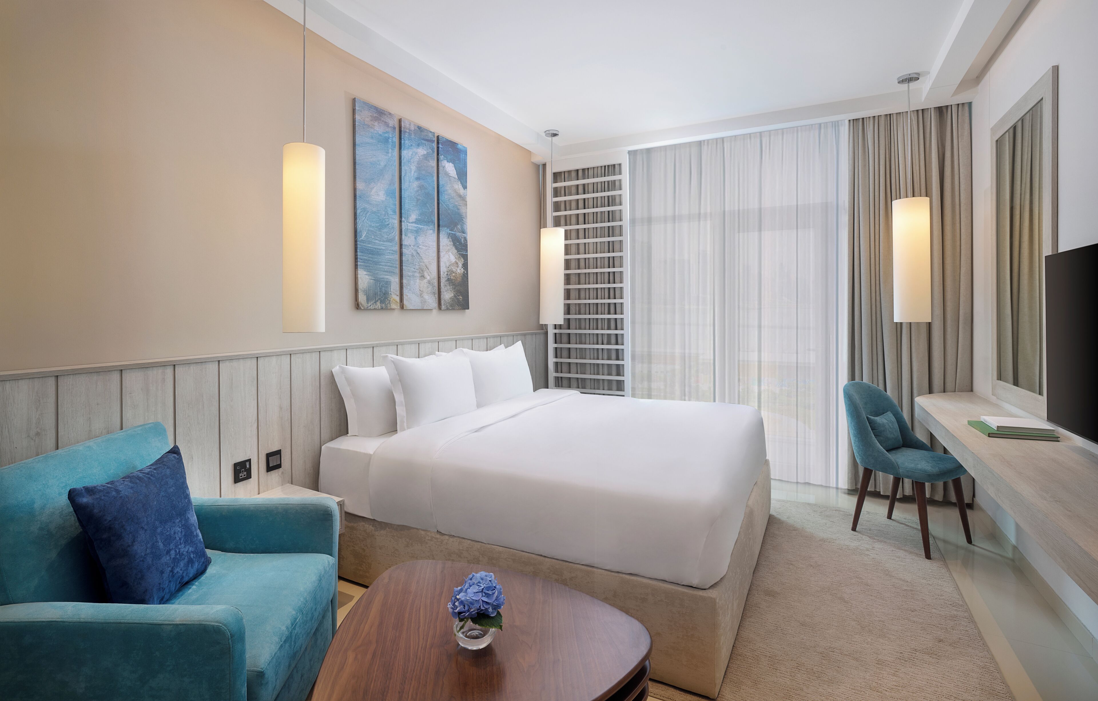 A hotel room with a large bed, a blue sofa, a desk with a chair, a TV, and a painting on the wall.
