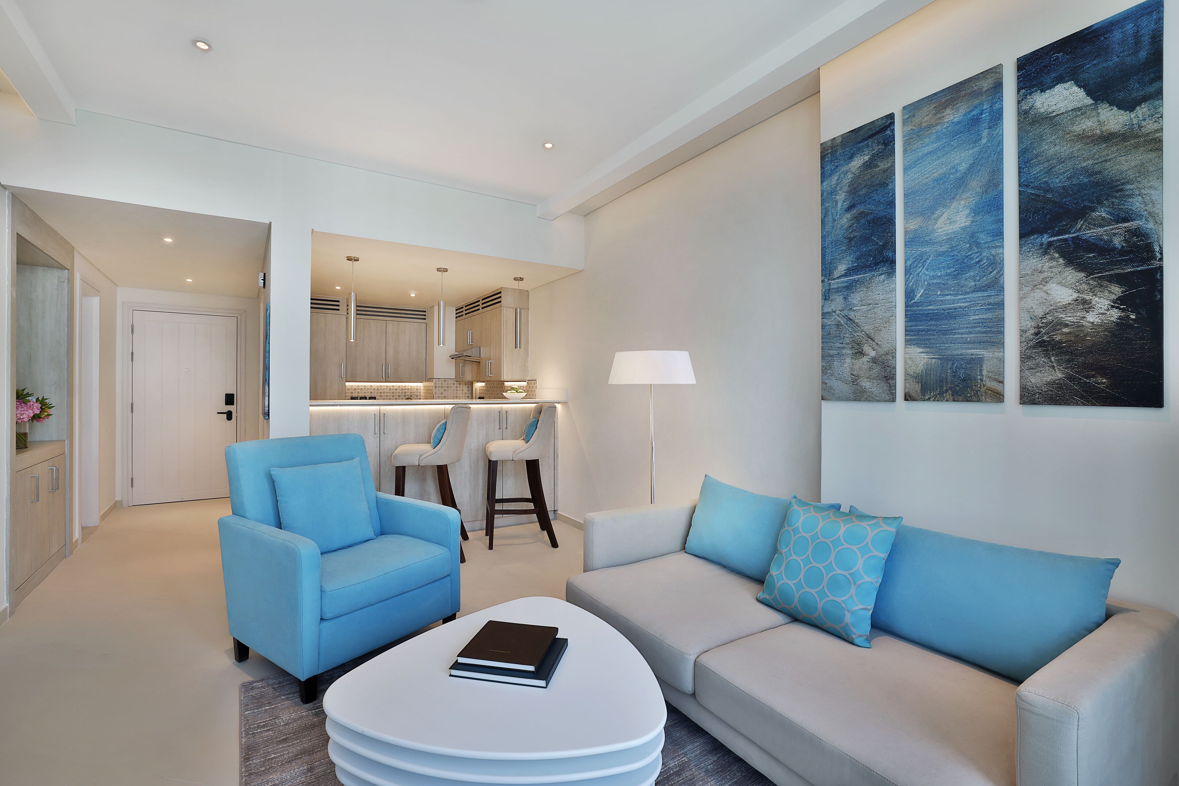 A modern living room with a sofa, armchair, and coffee table, a kitchen in the background, and a large abstract painting on the wall.