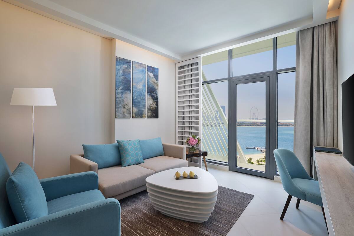 A modern living room with a sofa, armchairs, a coffee table, and a large window offering a view of the sea and a bridge.