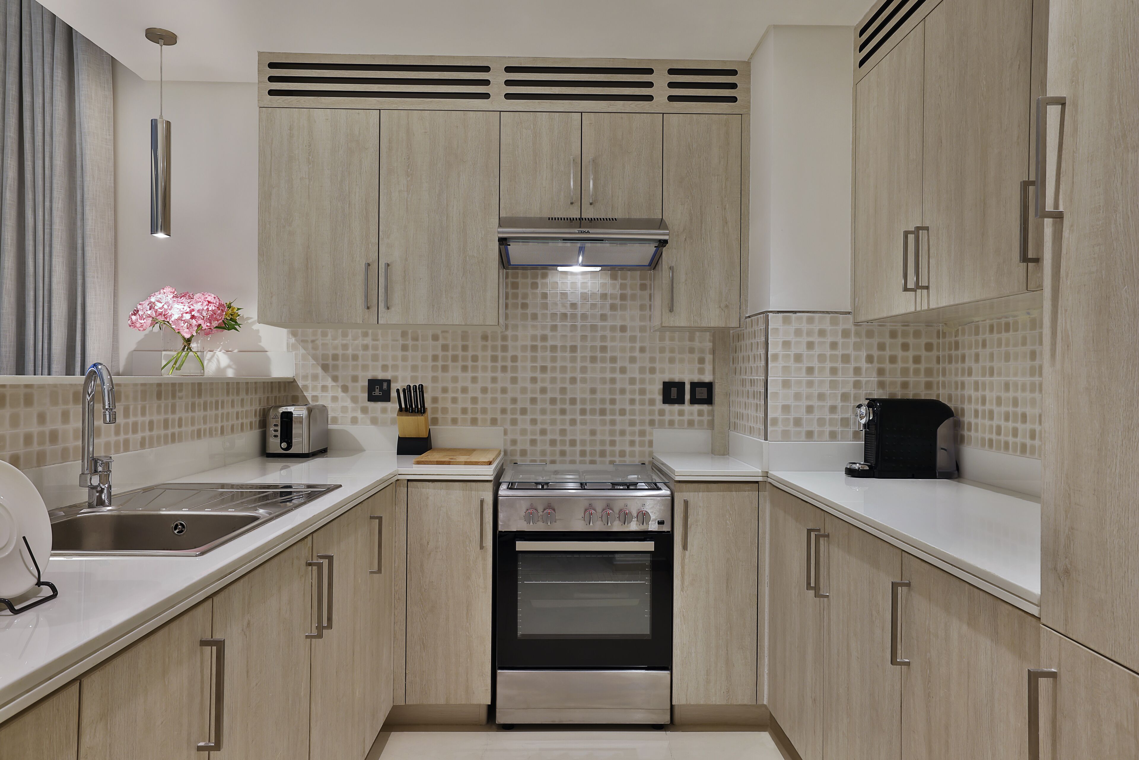 A modern kitchen with light wood cabinets, a stainless steel sink, a built-in oven, and a tiled backsplash.