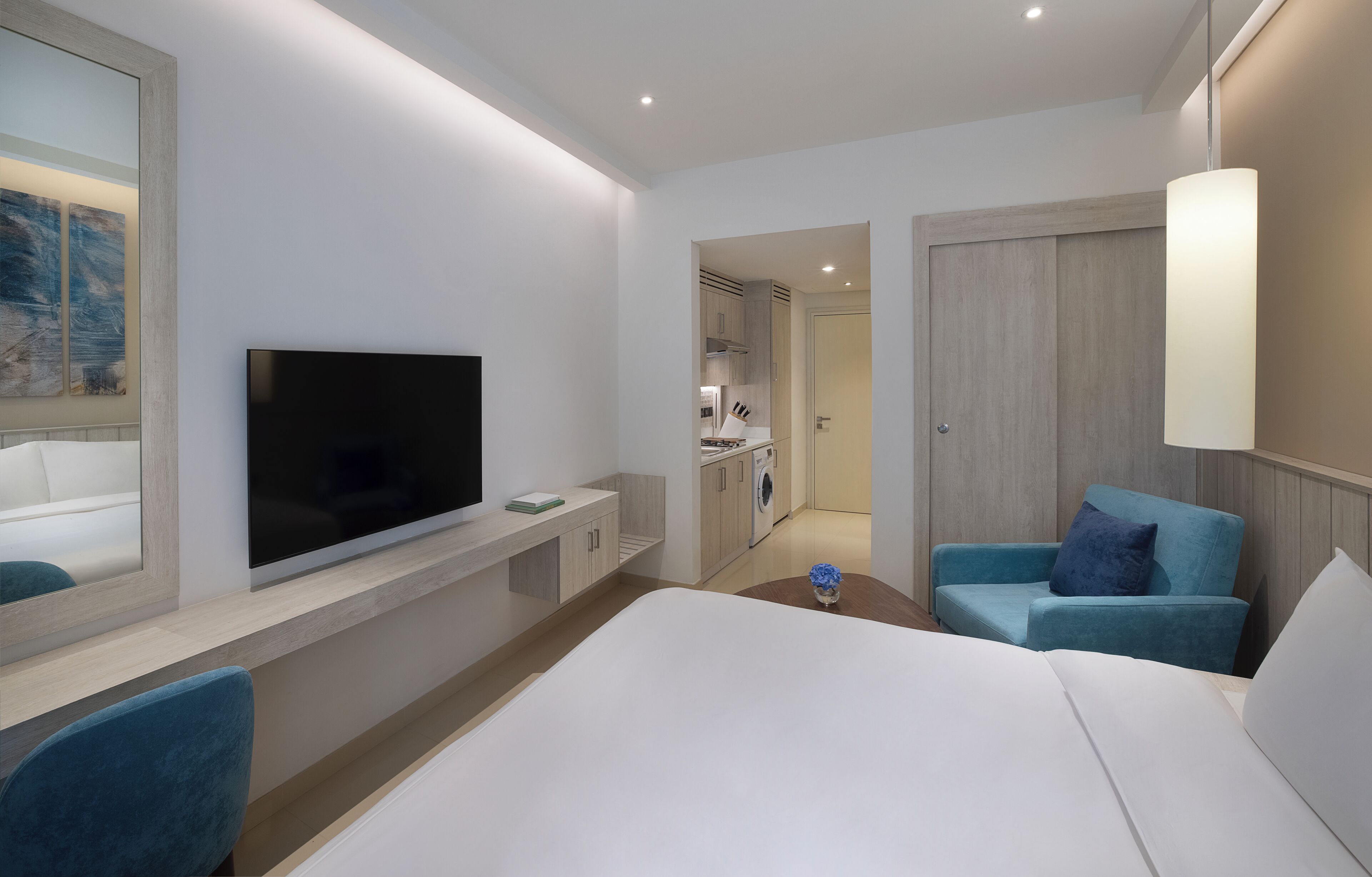 A hotel room with a large bed, a TV mounted on the wall, a seating area with a blue chair, and a small table with a vase.