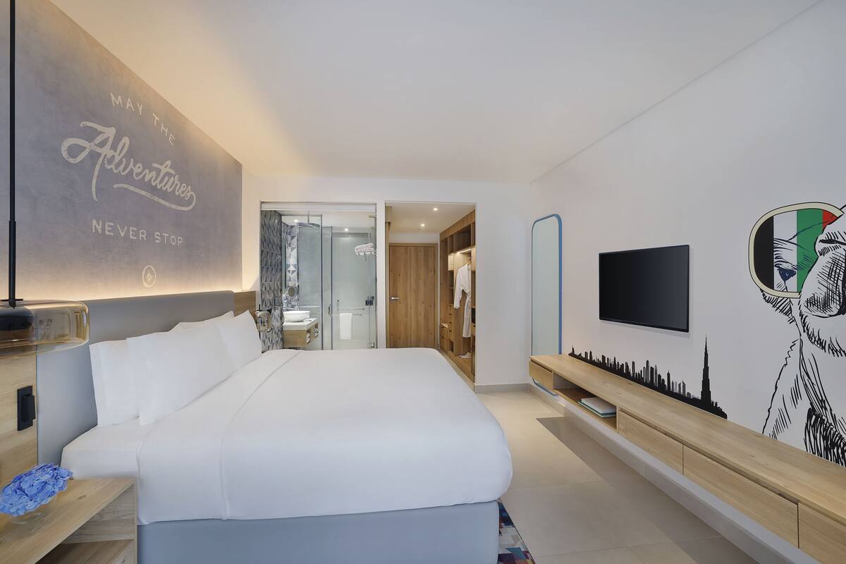 A modern hotel room with a large bed, a TV, and a city skyline mural on the wall.