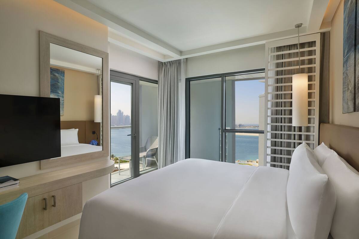 A hotel room with a large bed, a television, a desk with a chair, and a view of the sea through a sliding glass door.