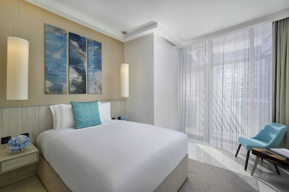 A hotel room with a large bed, a chair, a nightstand, and a window with sheer curtains.