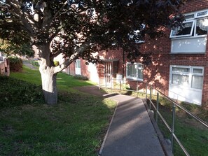 A pathway leading to a brick building with multiple windows, surrounded by greenery and a tree.