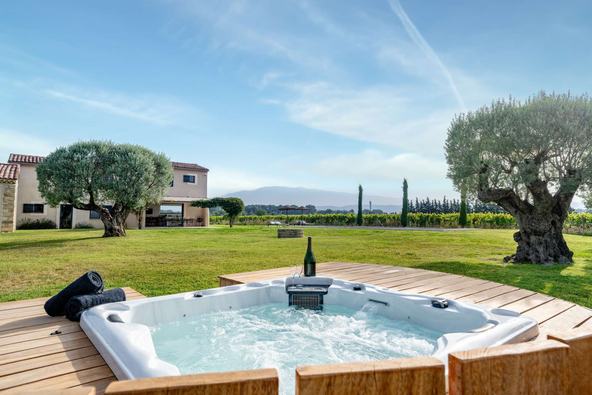 Een hottub op een houten terras, met uitzicht op een huis en een wijngaard op de achtergrond.