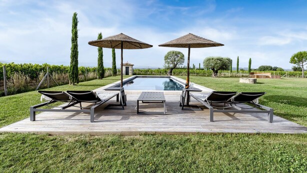 Zona de piscina con sillones de descanso y sombrillas, rodeada de vegetación y un cielo despejado.