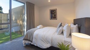 Een moderne slaapkamer met een groot bed, een bedlampje en uitzicht op een tuin door een glazen schuifdeur.