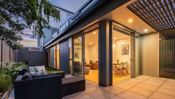 Een modern terras buiten, met een rieten bank, een eethoek en een glazen deur naar een binnenruimte.