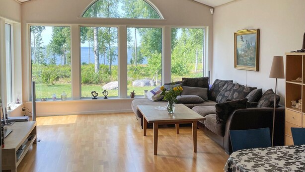 Un salon spacieux doté d’une grande fenêtre donnant sur un lac et des arbres, ainsi que d’un canapé, d’une table basse et d’une bibliothèque.
