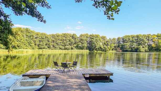 Un muelle de madera se extiende sobre un lago tranquilo, con una pequeña embarcación amarrada al final. Hay una mesa con sillas dispuestas en el muelle, y un cartel muestra el nombre 'Baden'.