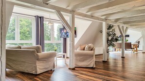 Un salón amplio con suelo de madera, dos sofás beige, una mesa de centro y un gran ventanal con cortinas.