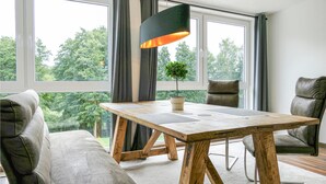 Un comedor moderno con una mesa de madera, dos sillas, una lámpara colgante y vistas a la vegetación a través de ventanas amplias.