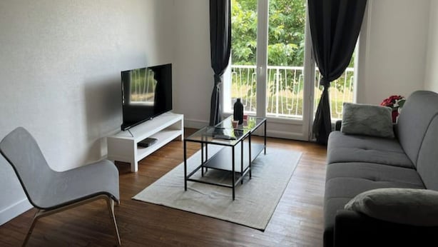 Uma sala de estar moderna com uma TV de tela plana em um suporte branco, uma mesa de centro de vidro, um sofá cinza e uma janela com cortinas pretas.