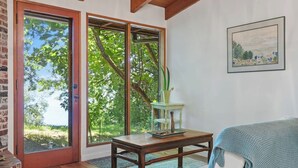 Une pièce dotée d’une grande porte-fenêtre coulissante donnant sur un jardin, d’une table en bois avec un vase, et d’un tableau encadré accroché au mur.