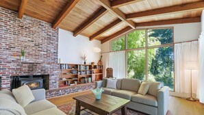Un salon chaleureux avec une cheminée en briques, un plafond en bois, une grande bibliothèque et une vue sur la verdure grâce à de grandes fenêtres.
