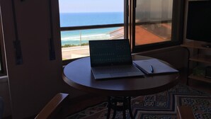 Een kamer met een laptop op een tafeltje, een stoel en uitzicht op de oceaan door een raam.