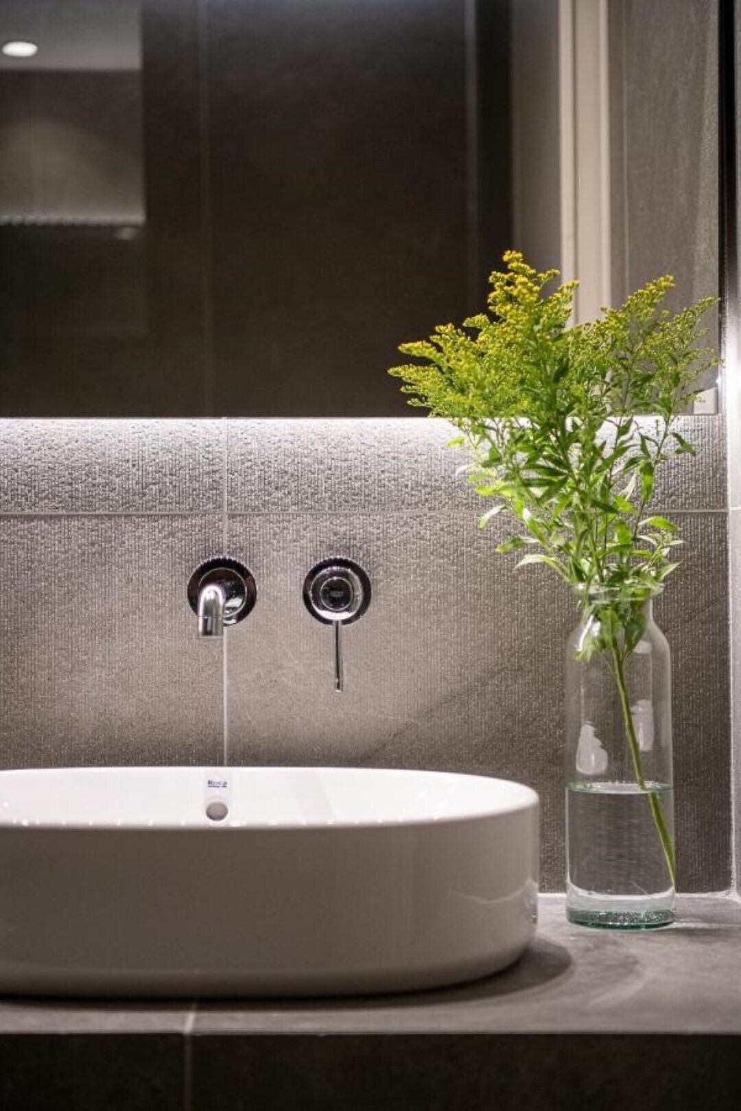 Ein Badezimmer mit einem runden weißen Waschbecken, zwei Wasserhähnen und einer Vase mit gelben Blumen auf der Ablagefläche.