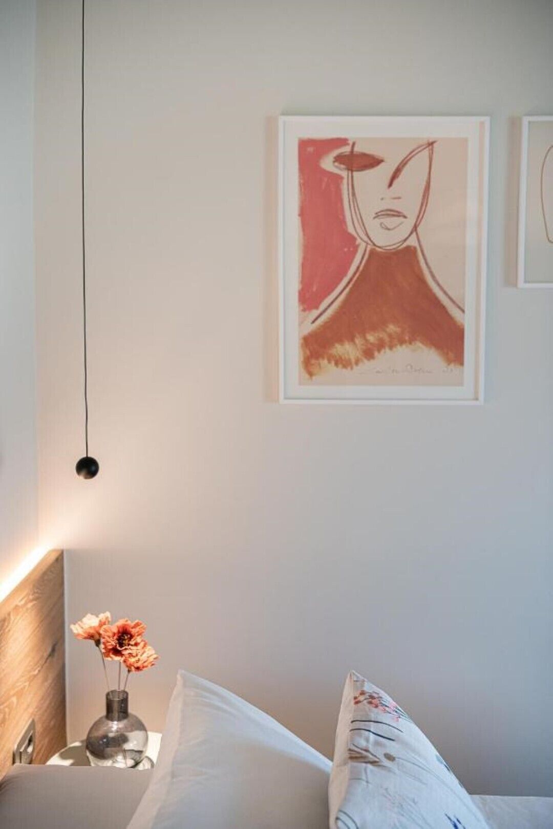 A bedroom with a bed, a bedside table with a vase of flowers, and a wall with framed artwork.