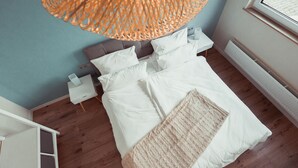 Ein Schlafzimmer mit einem großen Bett, weißer Bettwäsche, Holzboden, einer Pendelleuchte und einem Fenster mit Jalousien.