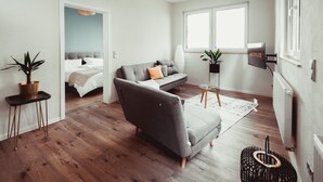 Ein modernes Wohnzimmer mit grauem Sofa, einem kleinen Tisch, einer Topfpflanze und einem an der Wand befestigten Fernseher.