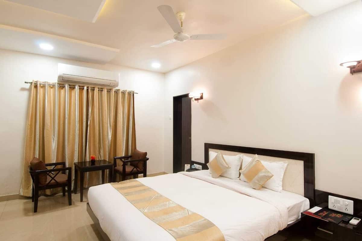 A hotel room with a large bed, a desk, two chairs, an air conditioning unit, a ceiling fan, and a bedside table with a phone and a book.