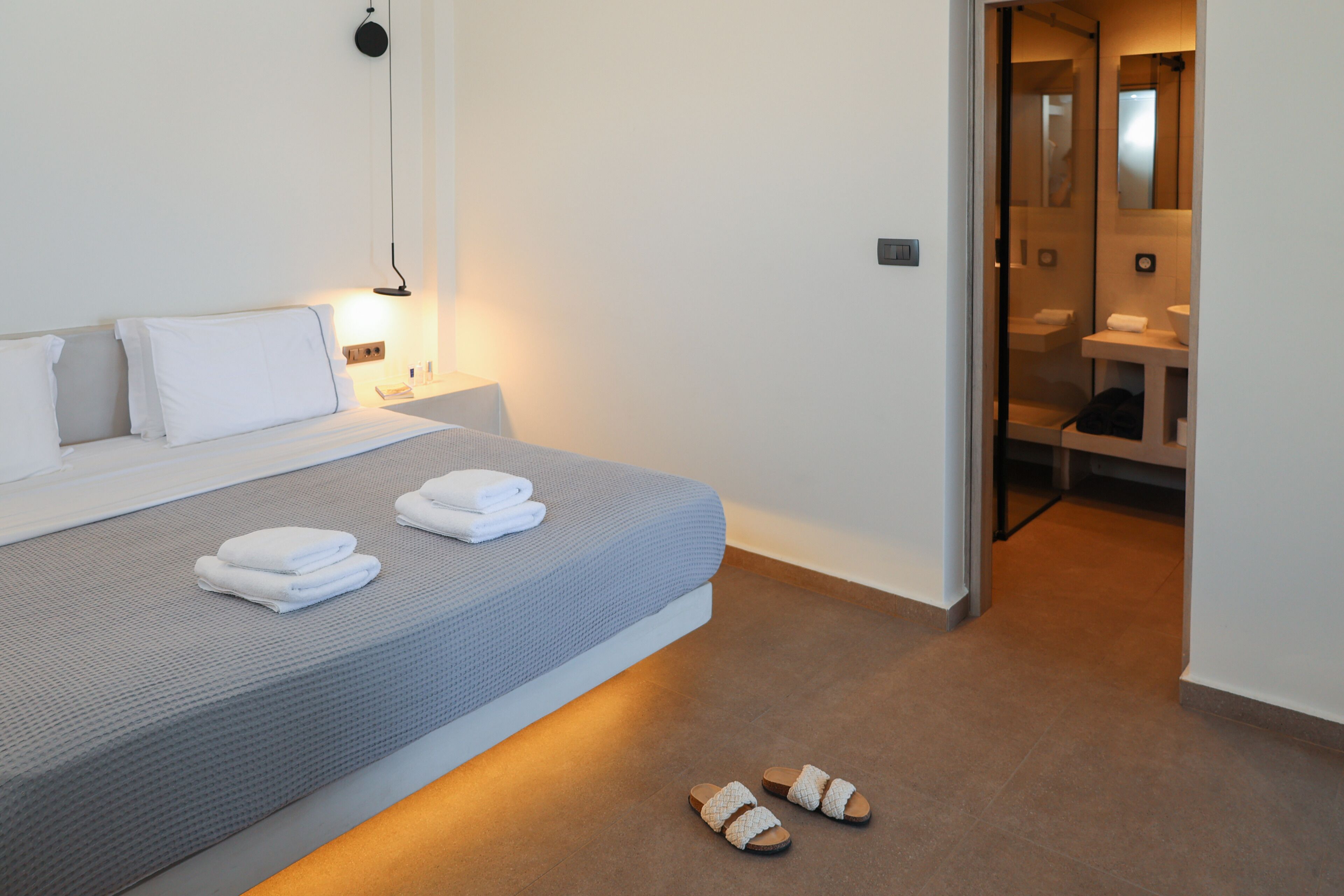 Una habitación de hotel moderna con una cama bien hecha, una mesita de noche con lámpara y un baño visible a través de una puerta abierta.