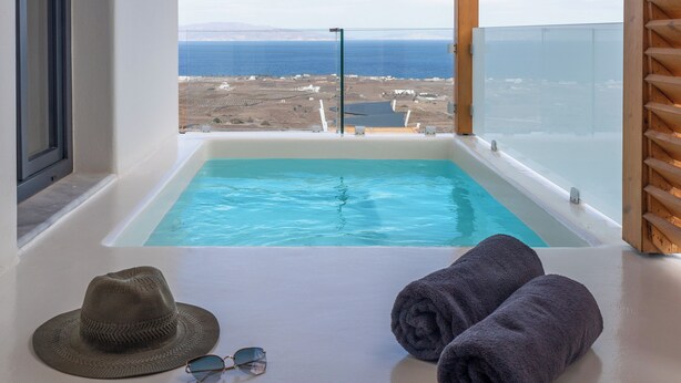 Una habitación moderna con techo de madera, un ventanal grande con vista a la playa y al océano, y una piscina pequeña con toallas y gafas de sol en el suelo.