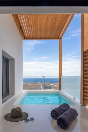 A modern room with a wooden ceiling, a large window overlooking a beach and ocean, and a small pool with towels and sunglasses on the floor. - North Wind Luxury Suites (Santorini)
