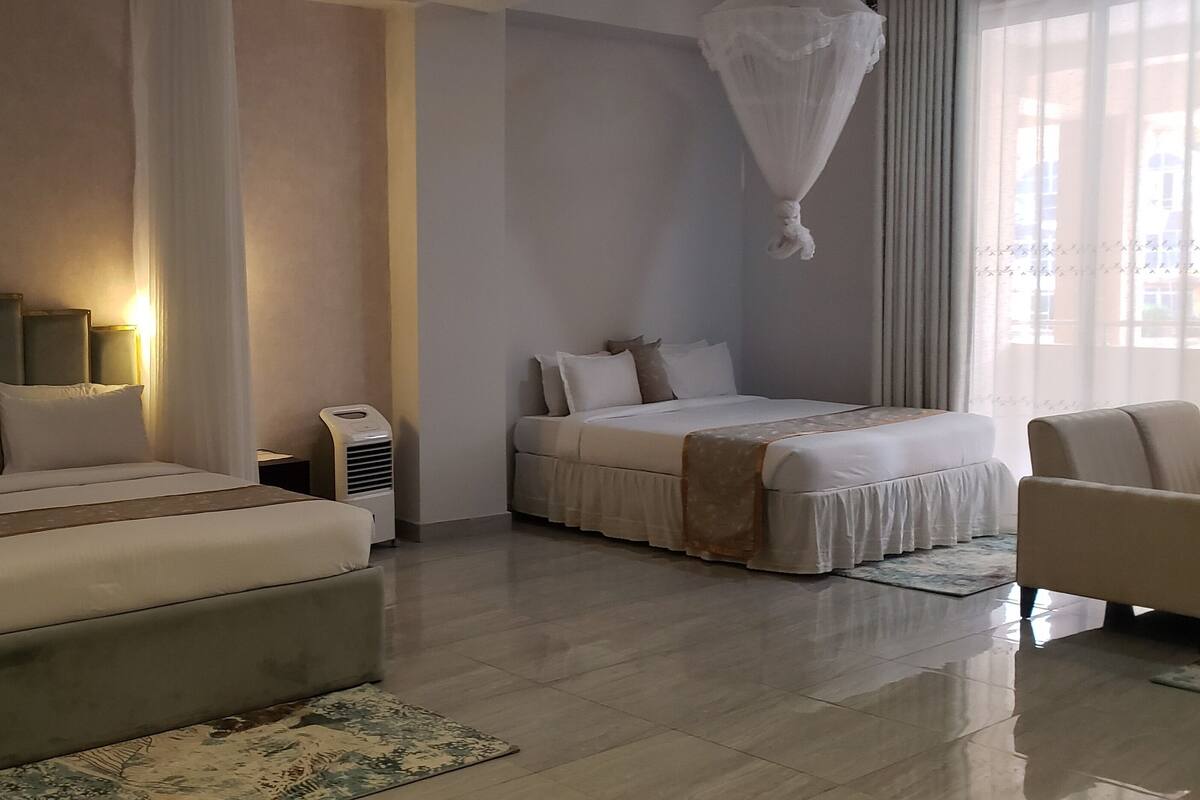 Una habitación de hotel con una cama grande, una cama más pequeña, un área de descanso con una silla, una mesita de noche y una ventana con cortinas.