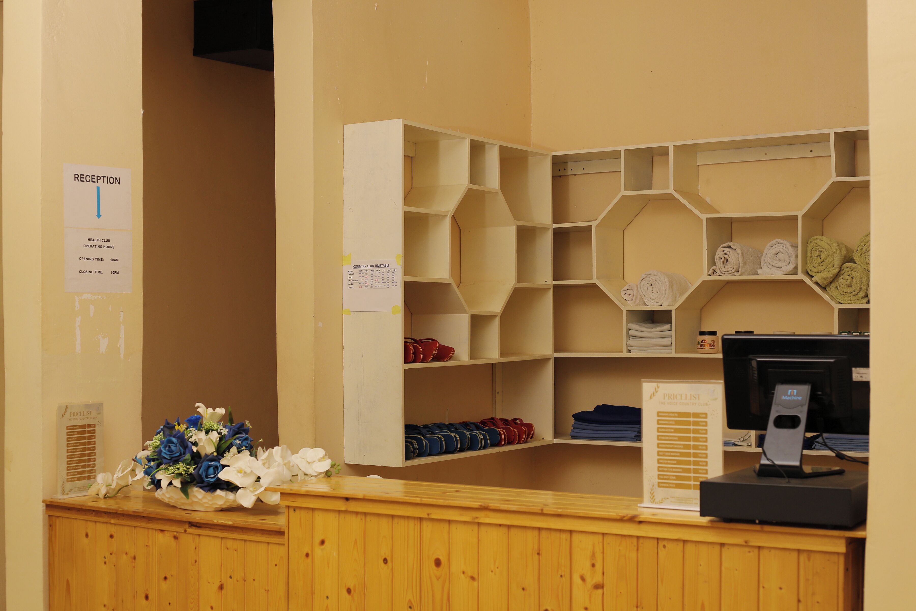 Un área de recepción con mostrador de madera, un letrero de recepción, un tablón de anuncios y un monitor de computadora.