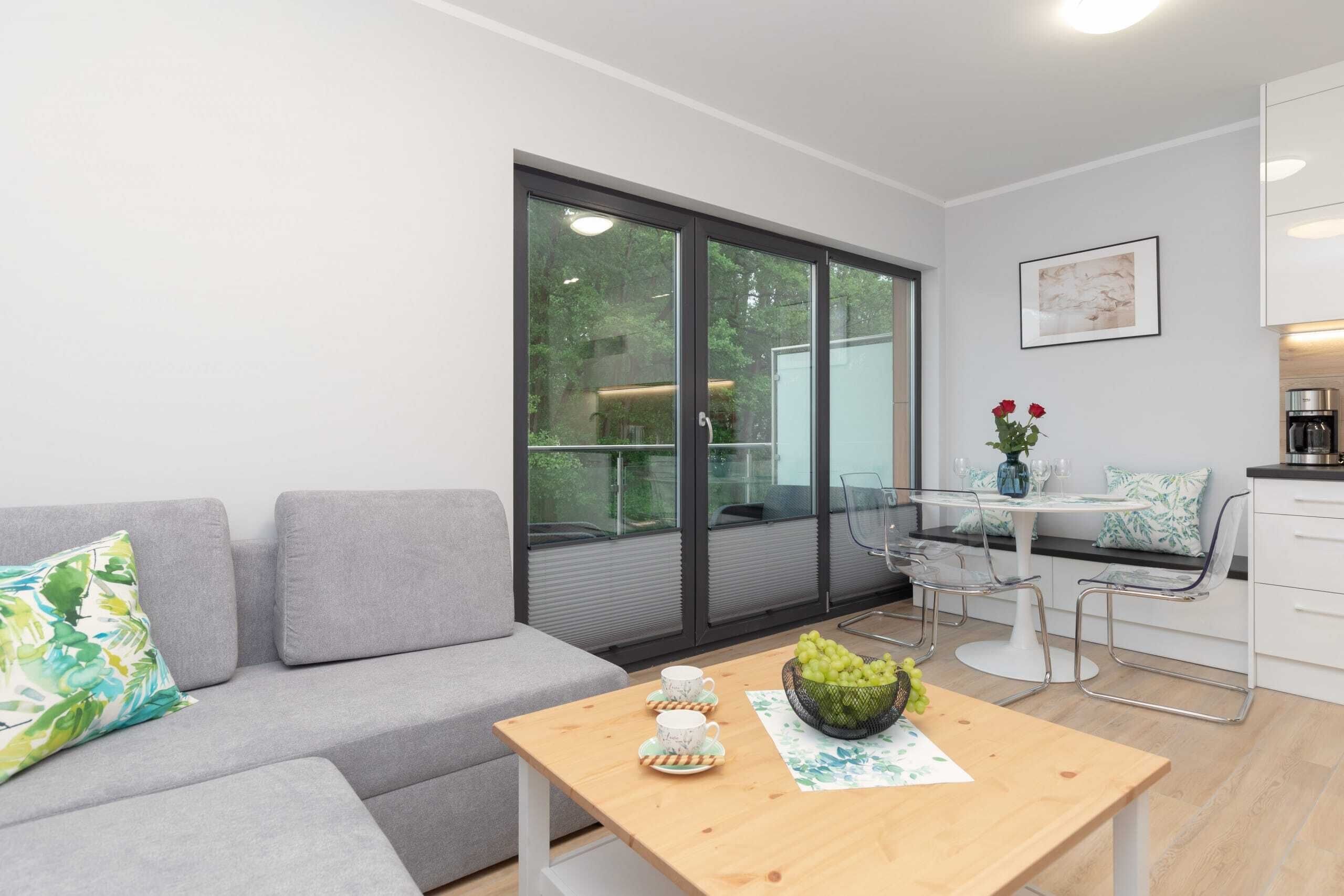 A modern living room with a grey sofa, a wooden coffee table with a vase and grapes, and a dining area with a white table and chairs.