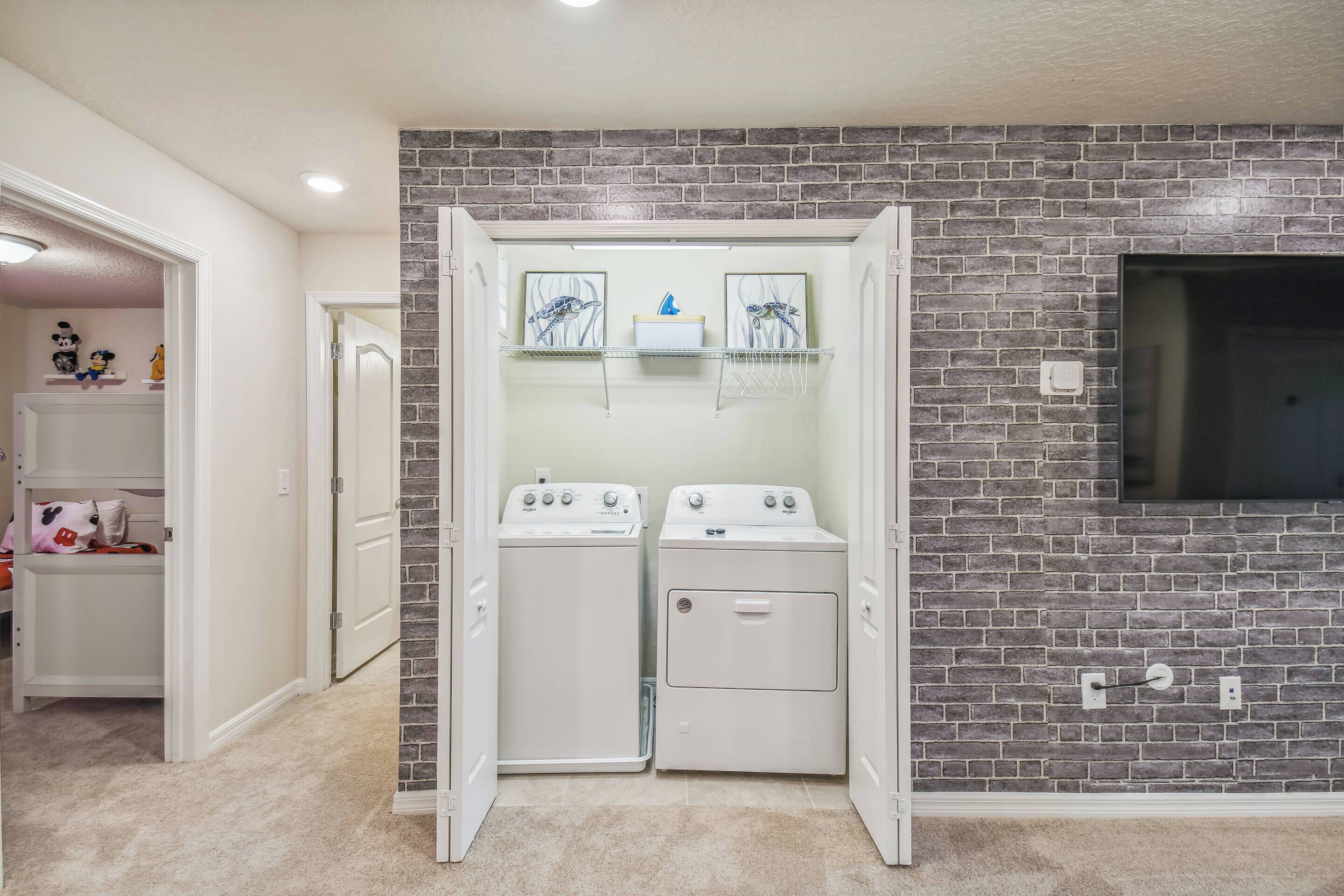 A laundry room with two white dryers, a brick wall, and a shelf with framed pictures.