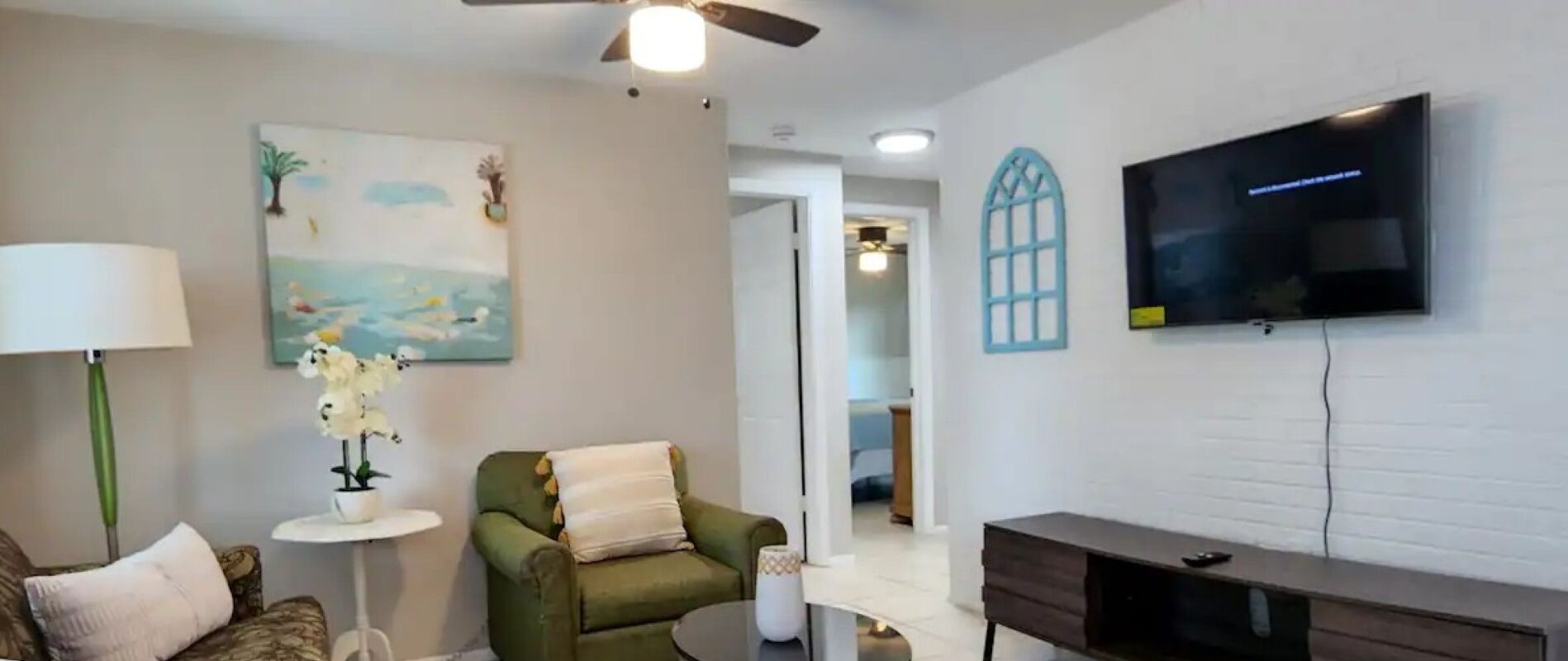 A living room with a green sofa, a small round table with a lamp, a painting on the wall, and a flat-screen TV mounted on the wall.
