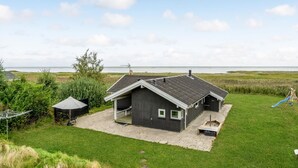 En lille, mørk hytte med skrå tag, overdækket terrasse og havudsigt.