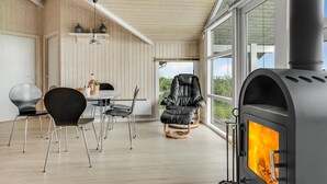 Et hyggeligt rum med pejs, et spiseområde med stole og bord, samt store vinduer der giver udsigt til naturen.