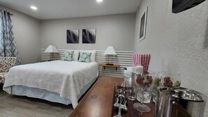 A bedroom with a bed, two bedside lamps, a wooden nightstand, and a table with glasses and a metal container. - #28 Little Belleview by Joyful Moments Vacation (Port Richey)