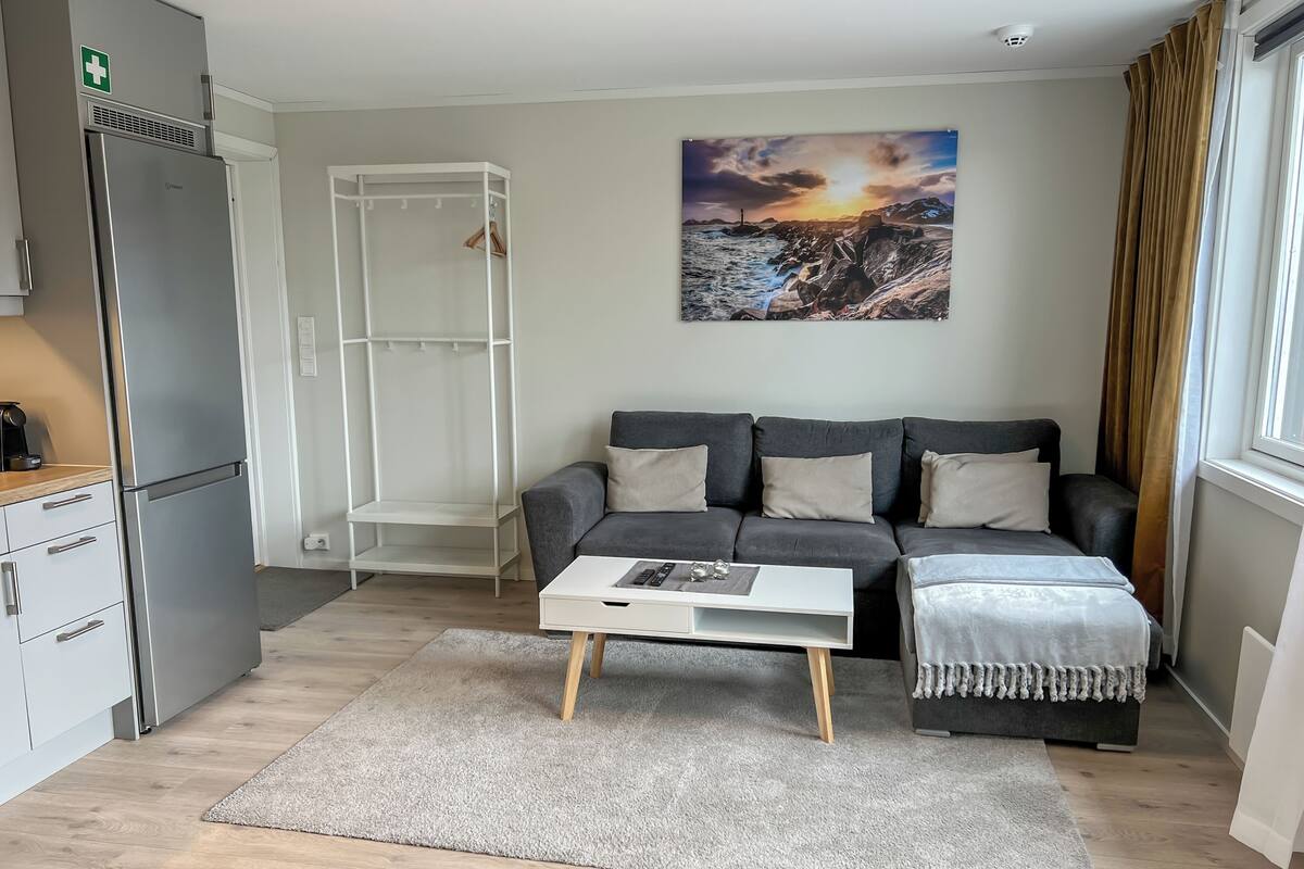 Ett modernt vardagsrum med en grå soffa, ett vitt soffbord, en tavla som föreställer en solnedgång och en köksdel med ett kylskåp.