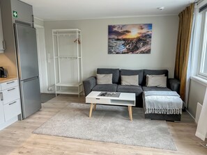 Ein modernes Wohnzimmer mit einem grauen Sofa, einem weißen Couchtisch, einem Gemälde von einem Sonnenuntergang und einer Küchenzeile mit einem Kühlschrank.