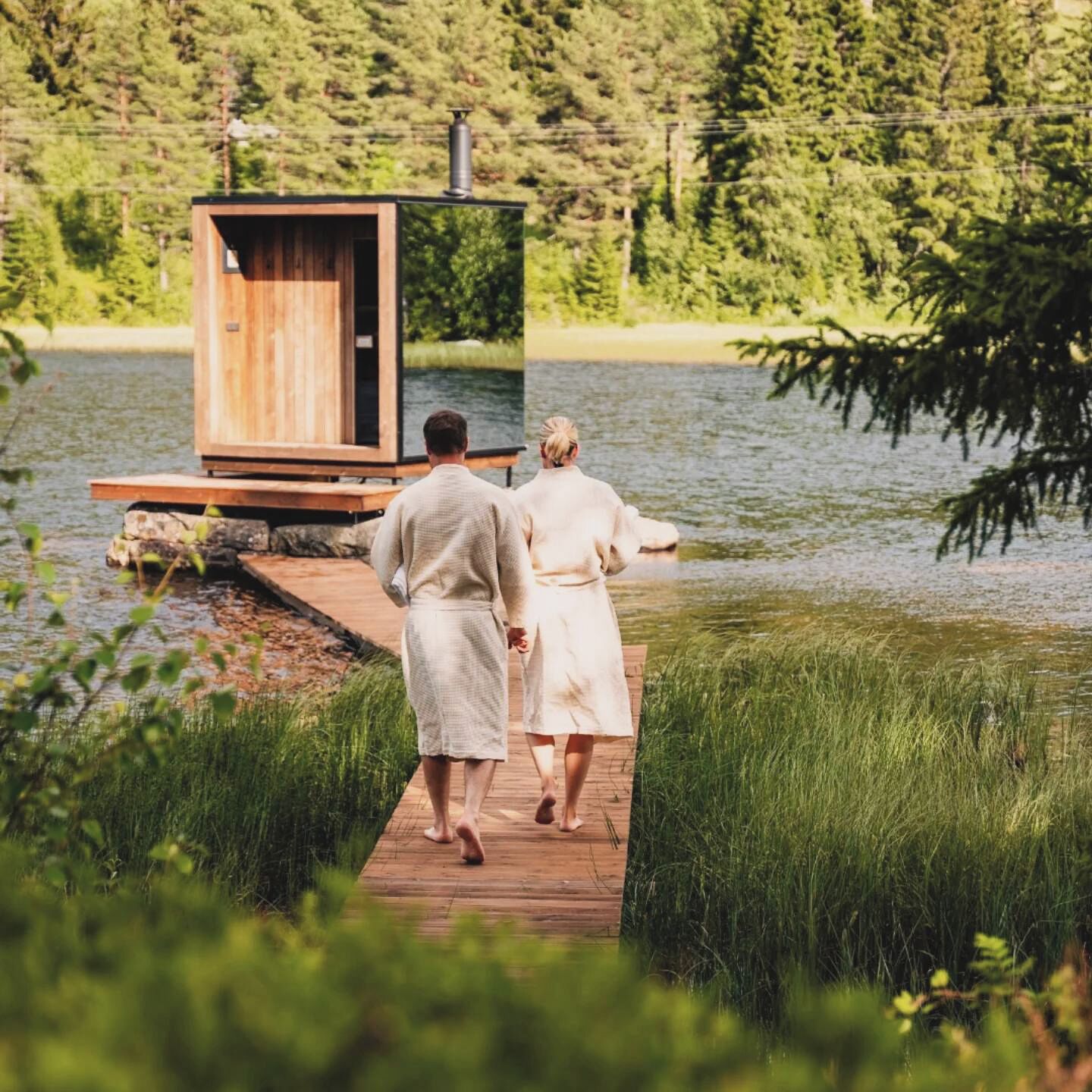 A wooden cabin with a small deck extending over a body of water, surrounded by lush greenery.