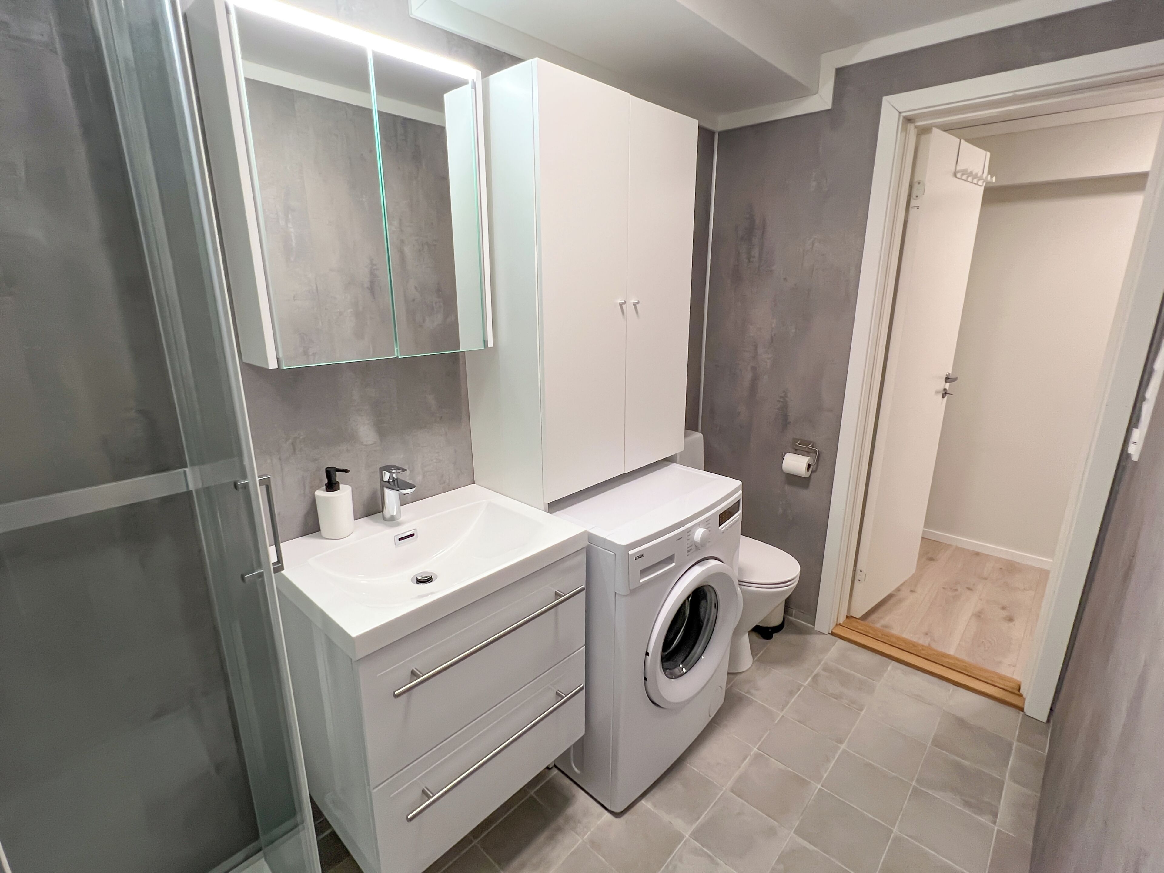 Un baño moderno con un lavamanos, espejo y armario blancos, una lavadora y un inodoro.