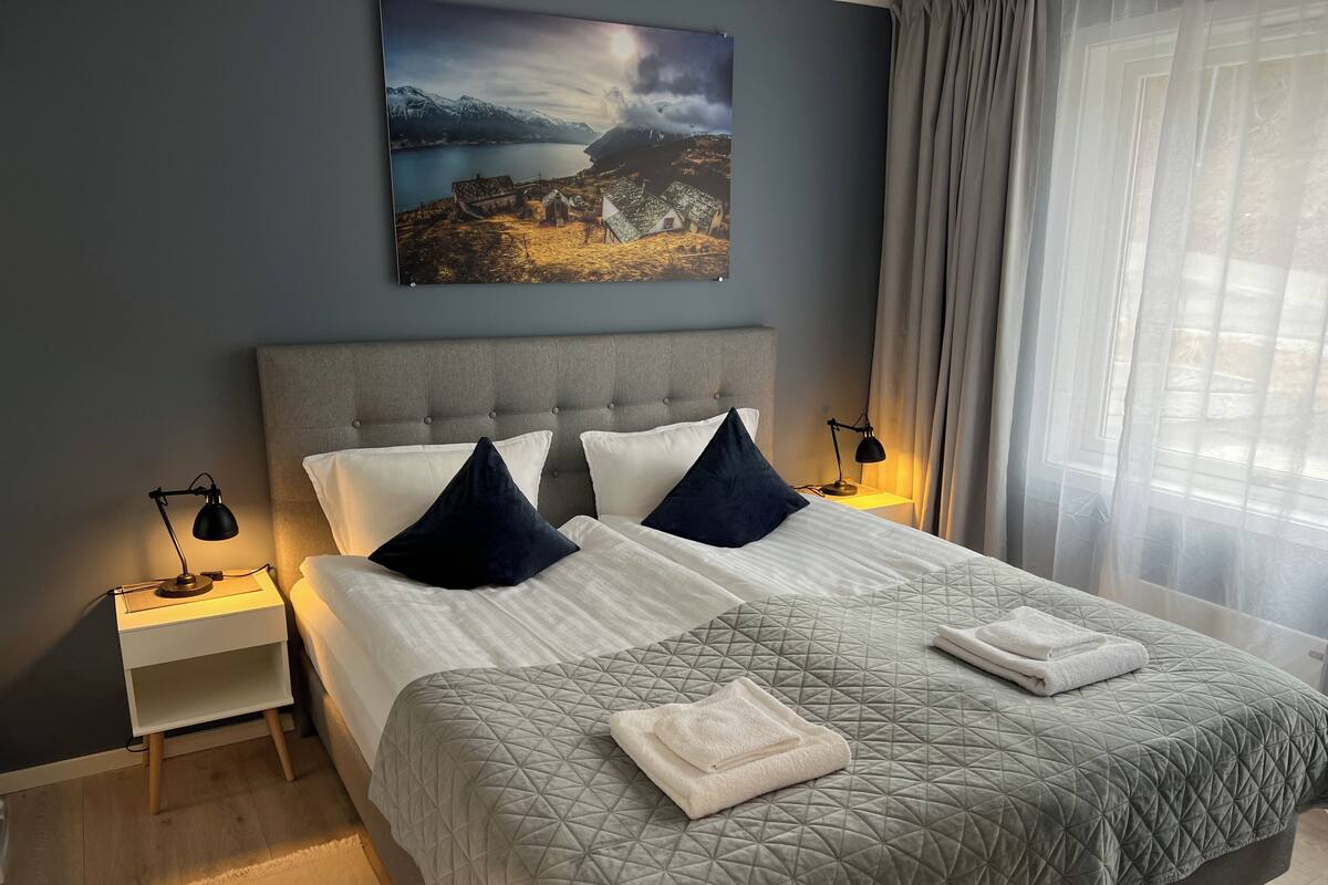 Ett hotellrum med en stor säng, två sängbord, ett litet bord, en tavla på väggen och ett fönster med gardiner.