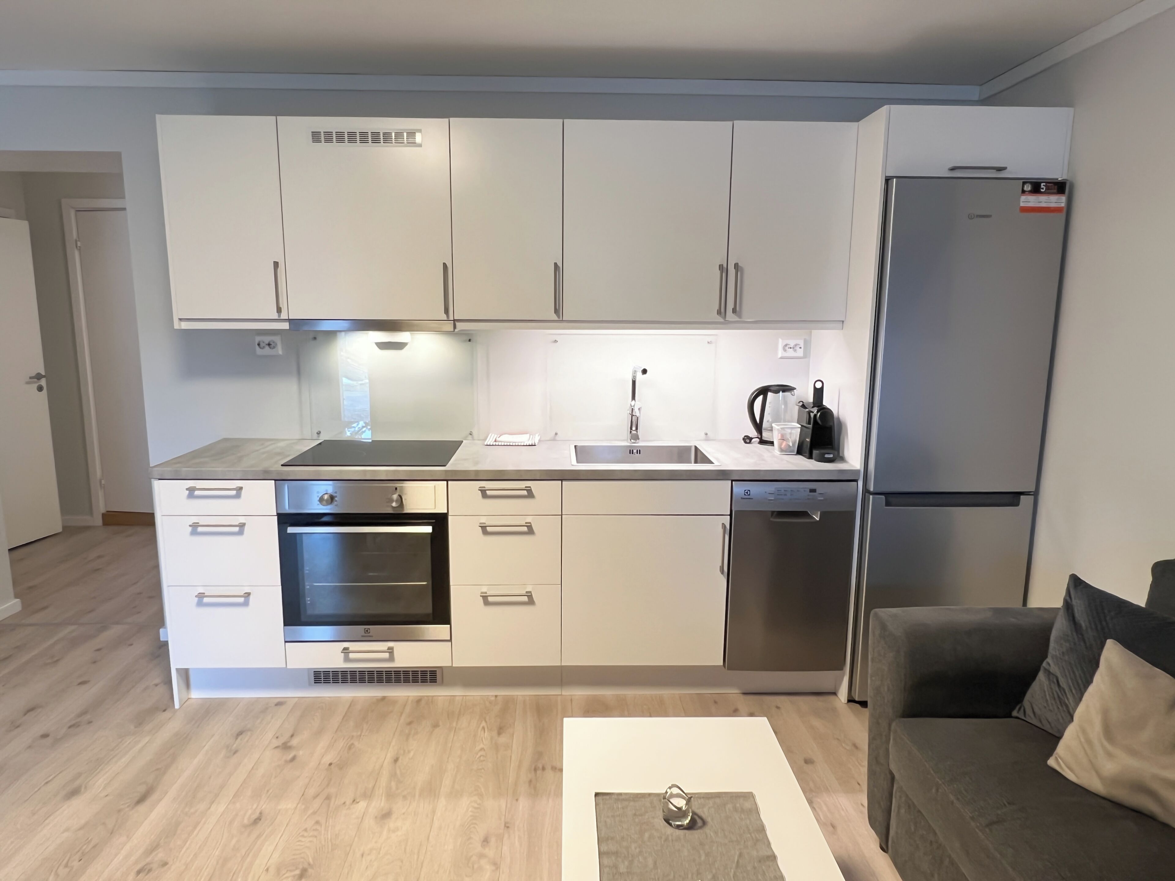 A modern kitchen with white cabinets, a stainless steel refrigerator, a built-in oven, and a coffee machine.