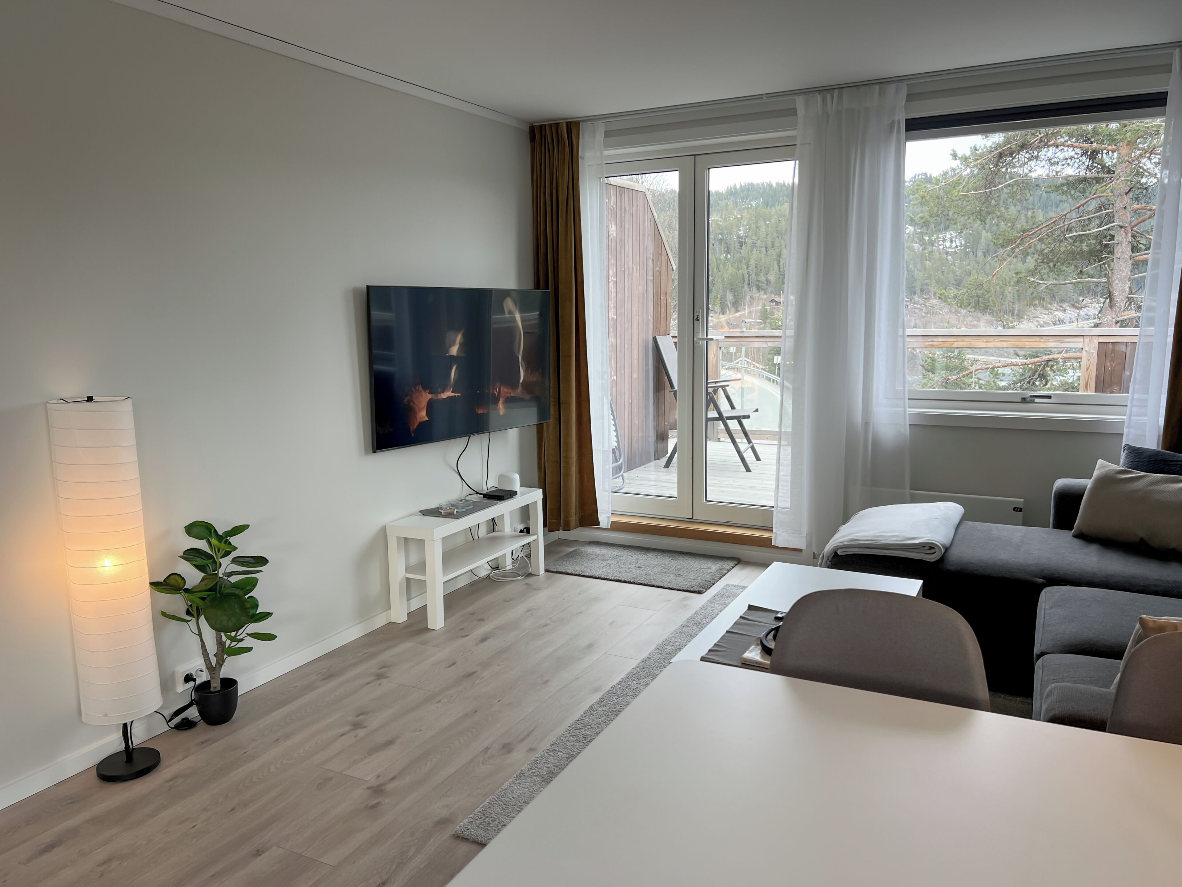 A modern living room with a flat-screen TV, a white console table, a grey sofa, and a large window with a view of trees.