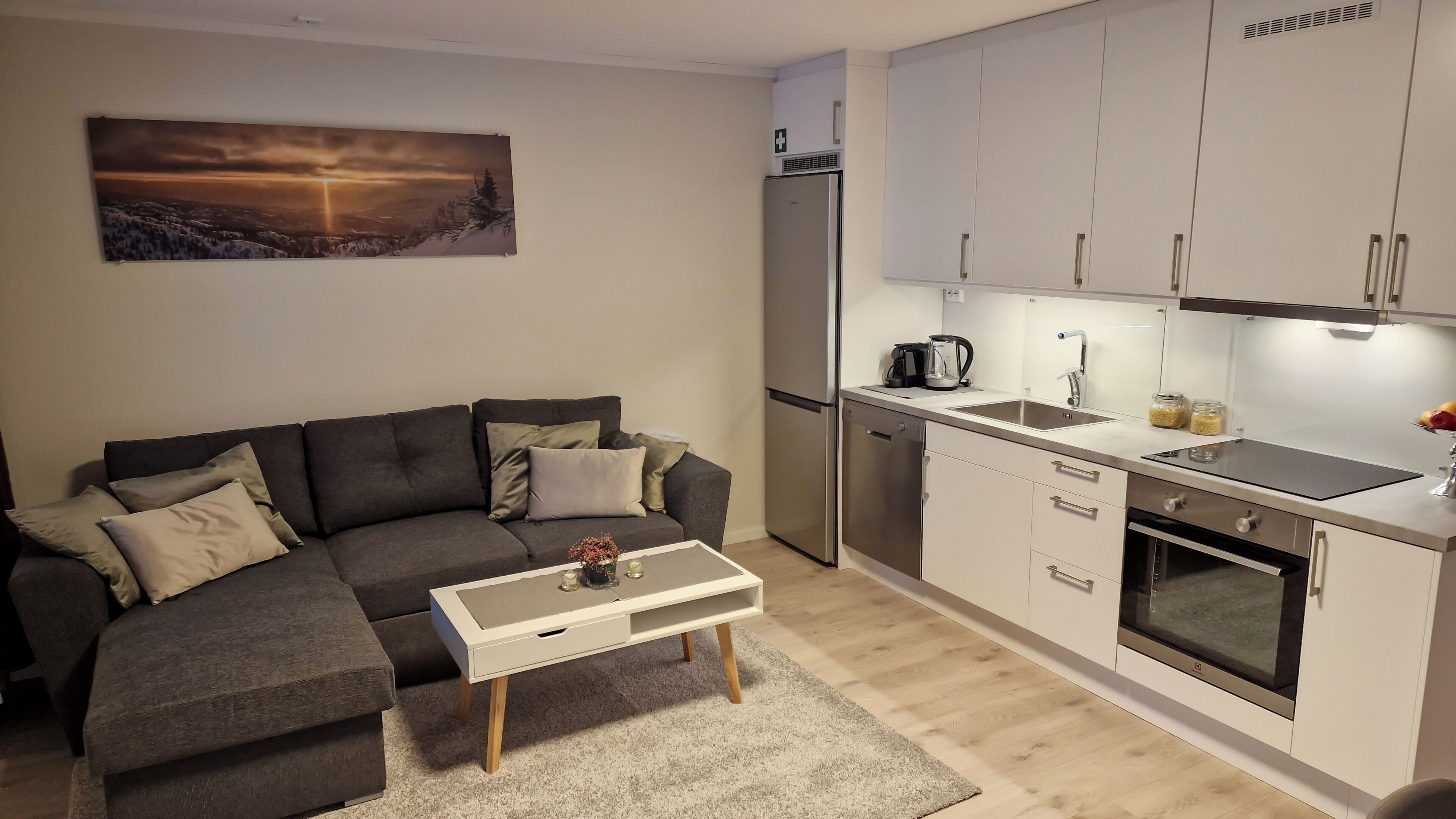 A modern living room with a grey sofa, a coffee table, and a kitchen area with white cabinets, a refrigerator, and a stove.