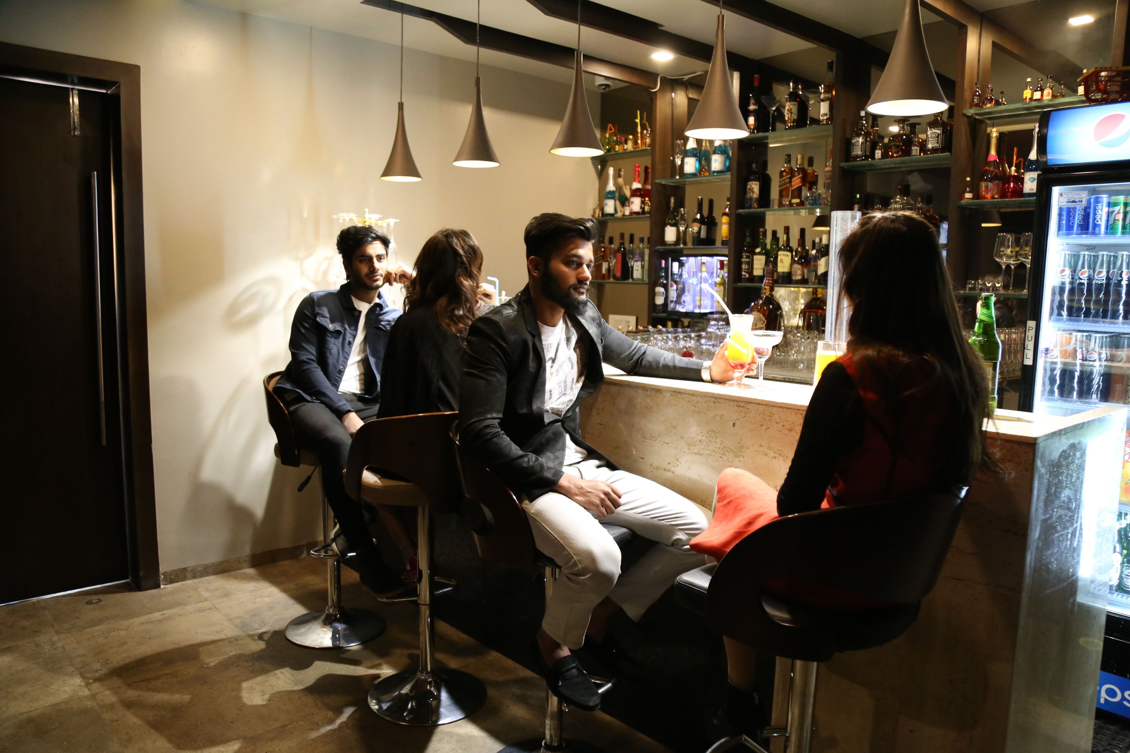 A bar area with multiple stools, a refrigerator stocked with beverages, and a variety of bottles on shelves.