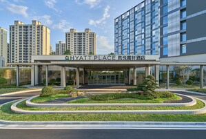 Una entrada moderna de hotel con un paseo techado, rodeada de jardines ajardinados y edificios residenciales altos al fondo.