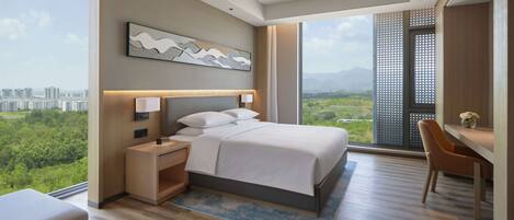 Habitación de hotel moderna con una cama grande, un escritorio y vistas a un paisaje urbano y zonas verdes.