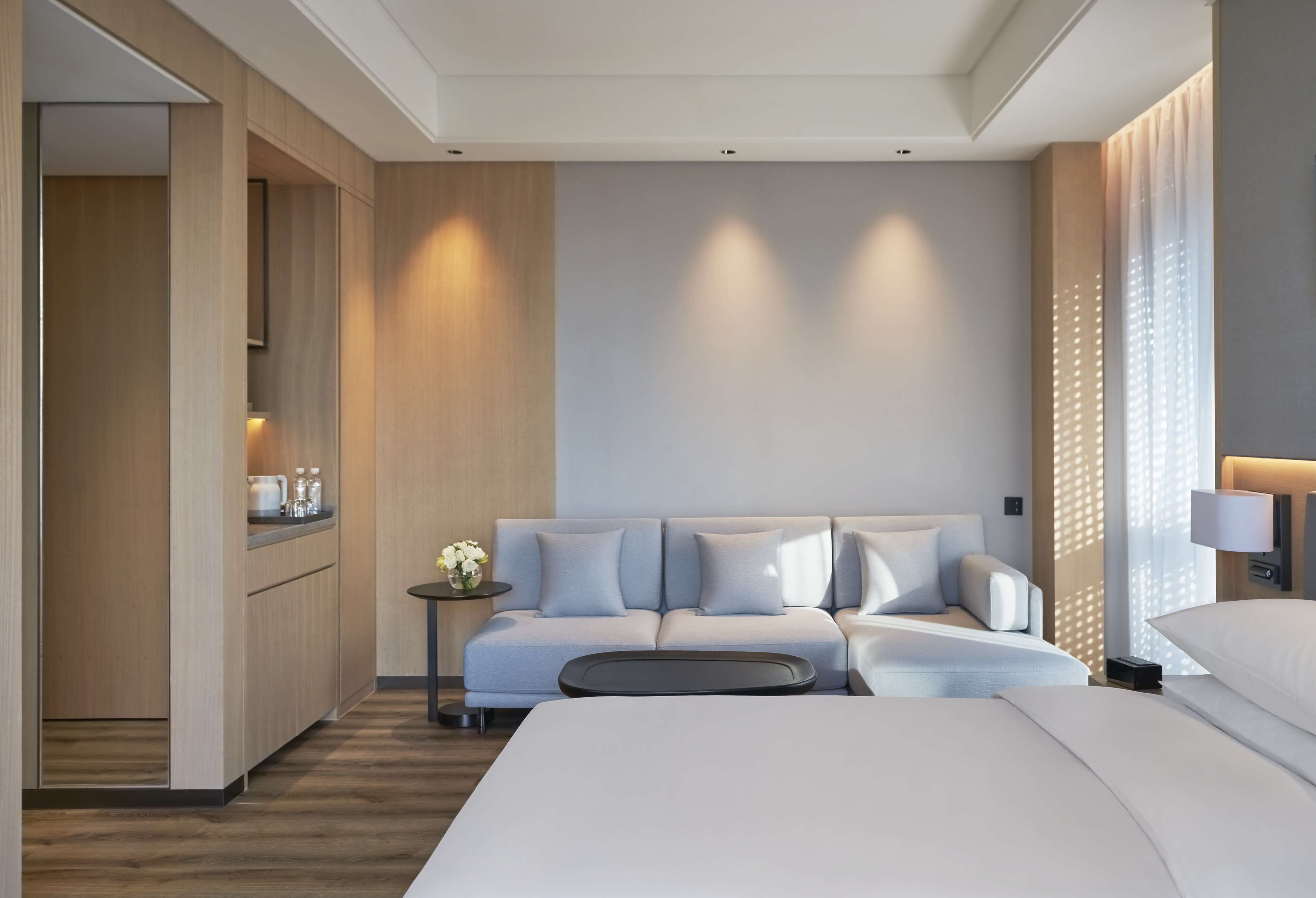 A modern hotel room with a large bed, a sofa, a small table with a vase of flowers, and a kitchenette area.