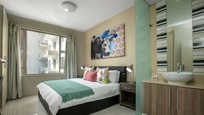 A modern bedroom with a large bed, a bedside table, a nightstand, a bathroom with a sink and mirror, and a window with curtains. - The Tyrwhitt Unit 408 (Johannesburg)